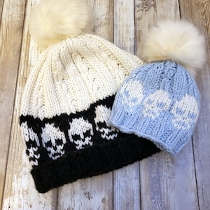 May include: Two knitted hats with white pom poms. The larger hat is white with a black skull pattern and the smaller hat is light blue with a white skull pattern.