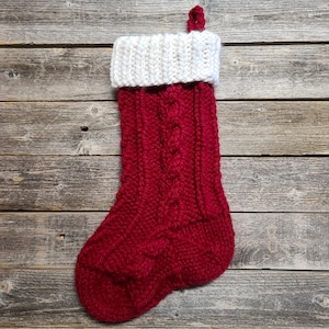 May include: A red and white knitted Christmas stocking with a cable knit design. The stocking has a white cuff and a loop for hanging. The background is a rustic wooden surface.