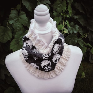 May include: A white and black knitted cowl scarf with a repeating pattern, displayed on a white mannequin. The scarf has a ruffled edge and is made of soft yarn. The background features green foliage.