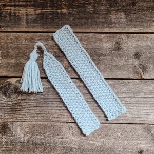 May include: Two light blue, knitted bookmarks with a matching tassel, resting on a rustic wooden surface. The bookmarks are rectangular, with a textured stitch pattern. The tassel is made of soft, light blue yarn.