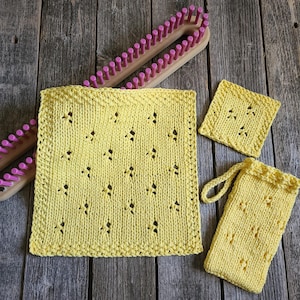 May include: A yellow knitted square washcloth, a smaller yellow knitted square, and a yellow knitted pouch with a loop, all featuring a repeating eyelet pattern, displayed alongside a long wooden knitting loom with pink pegs.