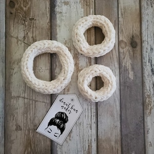 Op de afbeelding: Twee witte gebreide donut-broodjesmakers op een houten achtergrond. De grotere donut-broodjesmaker bevindt zich in het midden van de afbeelding. De kleinere donut-broodjesmaker bevindt zich rechts van de grotere. Een wit label met zwarte tekst dat "donut bun maker" zegt, bevindt zich in de linkerbenedenhoek van de afbeelding.