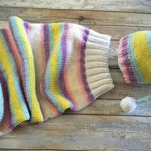 Puede incluir: Un gorro de bebé tejido y una envoltura a juego en un patrón de rayas de arcoíris con un pompón blanco en el gorro.