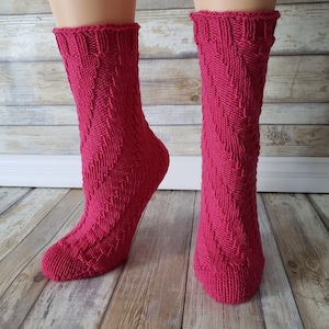 Op de afbeelding: Een paar roze gebreide sokken met een chevron-patroon. De sokken worden getoond op een paar benen, met de voeten rustend op een houten vloer.