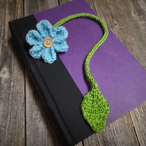 May include: A crocheted bookmark featuring a blue flower and green leaf. The bookmark is attached to a book with a purple cover.