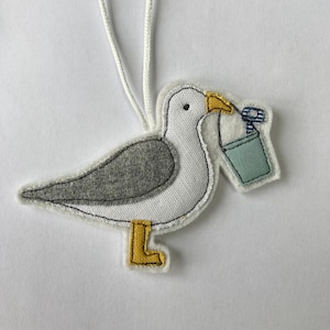 May include: A grey and white seagull ornament with yellow feet holding a blue bucket with a white and blue striped handle. The ornament has a white loop for hanging.