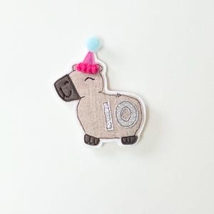 May include: A whimsical, light brown capybara-shaped patch with a pink party hat and a blue pom-pom. The number "10" is embroidered on the capybara's body with silver glitter. The patch has a white border.