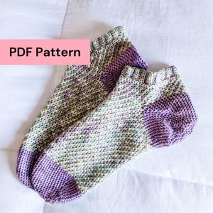 May include: A pair of knitted socks with a textured stitch pattern. The socks feature a colour palette of green, purple, and white. The heels and toes are a solid purple. The text "PDF Pattern" is displayed on a pink background.