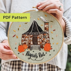 May include: Embroidered hoop art depicting Hagrid's Hut with pumpkins and autumn leaves. The design shows a stone cottage with a black roof and the text "Hagrid's Hut". The background is a light green fabric, and the hoop is gold.