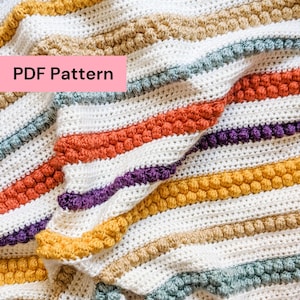 Op de afbeelding: Een handgemaakte gehaakte deken met een gestreept patroon. De deken heeft afwisselende rijen witte en kleurrijke strepen, waaronder oranje, paars, geel en bruin. De tekst "PDF Pattern" is zichtbaar op de afbeelding.