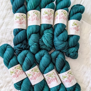 May include: Nine skeins of deep teal yarn arranged in a cluster. Each skein features a white paper label with a botanical illustration of red berries and green leaves, and the text Woolberry Fiber Co. printed in a cursive script font.