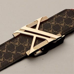 Op de afbeelding: Een bruine leren riem met een goudkleurige gesp. De gesp heeft de vorm van een gestileerde letter "A". De riem heeft een herhalend patroon van een logo. De gesp heeft een getextureerd oppervlak.