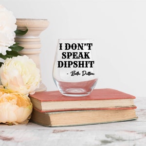 May include: A clear stemless wine glass with the text "I DON'T SPEAK DIPSHIT - Beth Dutton" in black. The glass is on top of two vintage books with red covers. Decorative flowers and a wooden candle holder are in the background.