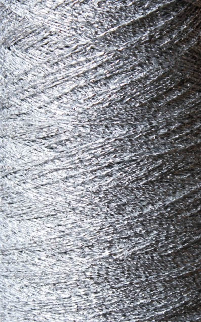 May include: Close-up of a spool of silver yarn. The yarn is tightly wound, showing a textured surface with a metallic sheen. The yarn appears to be made of a shiny, reflective material, ideal for crafting projects.