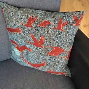 Puede incluir: Una almohada de sofá azul turquesa con un patrón de pájaros rojos volando sobre un fondo blanco. Los pájaros son estilizados y tienen un diseño geométrico.