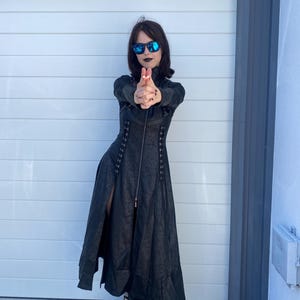 May include: A black long coat with a textured pattern, a front zipper, and a high collar. The coat is paired with black platform boots with silver buckles. The person is wearing sunglasses.