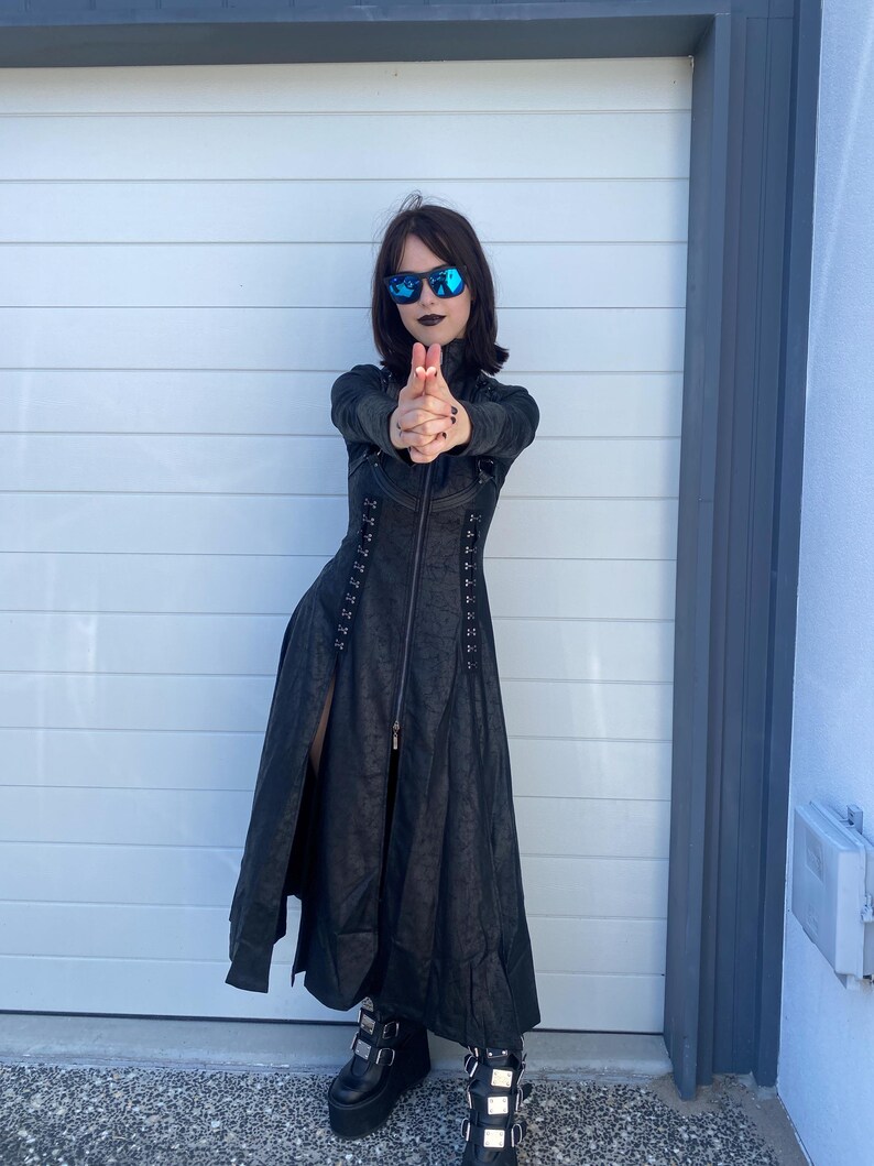 May include: A black long coat with a textured pattern, a front zipper, and a high collar. The coat is paired with black platform boots with silver buckles. The person is wearing sunglasses.