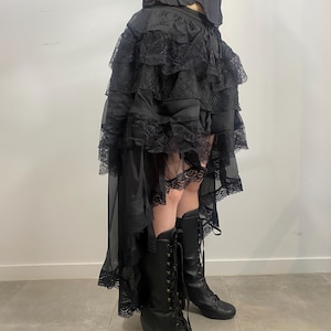 May include: Black layered skirt with lace trim and a high-low hemline. The skirt is paired with a black corset top and black lace-up boots with a block heel. The outfit has a gothic or Victorian aesthetic.