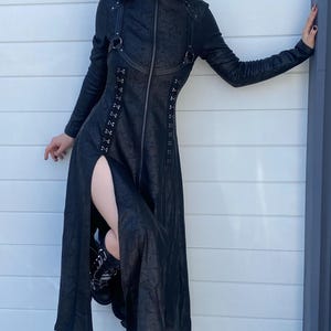 May include: Black long coat with a high collar and a front zipper. The coat features a thigh-high slit and is paired with platform boots. The outfit is completed with black sunglasses.