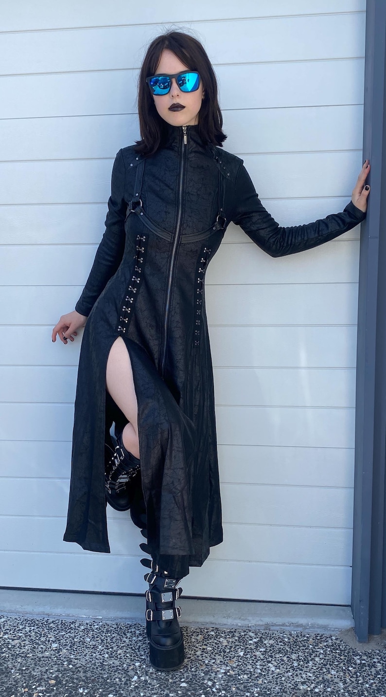 May include: Black long coat with a high collar and a front zipper. The coat features a thigh-high slit and is paired with platform boots. The outfit is completed with black sunglasses.