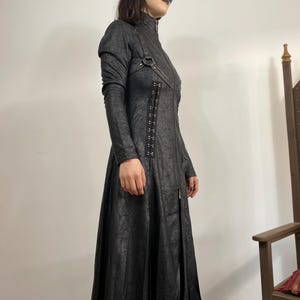 May include: A long black coat with a high collar, long sleeves, and a full-length zipper. The coat features decorative metal hardware and a textured pattern. The coat is a dark, gothic style.