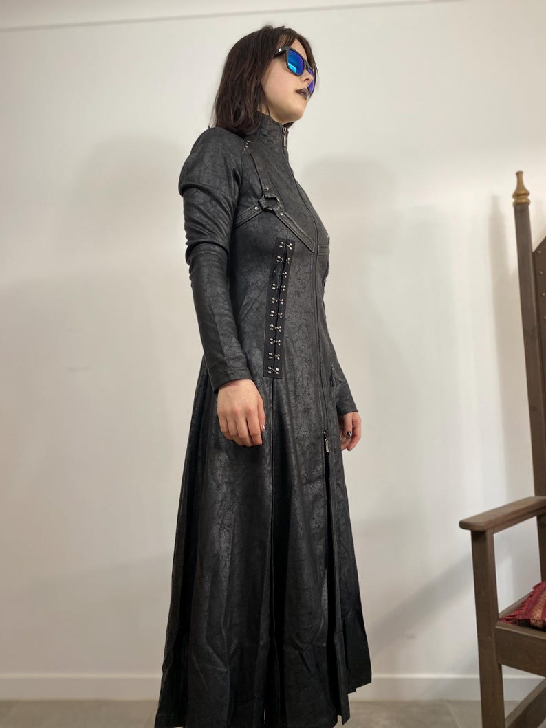 May include: A long black coat with a high collar, long sleeves, and a full-length zipper. The coat features decorative metal hardware and a textured pattern. The coat is a dark, gothic style.