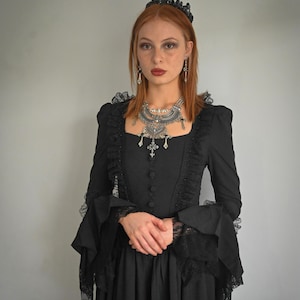 Op de afbeelding: Een vrouw in een zwarte jurk met kantdetails en een zwarte kroon. Ze draagt een zilveren ketting met een kruis hanger en zilveren oorbellen.