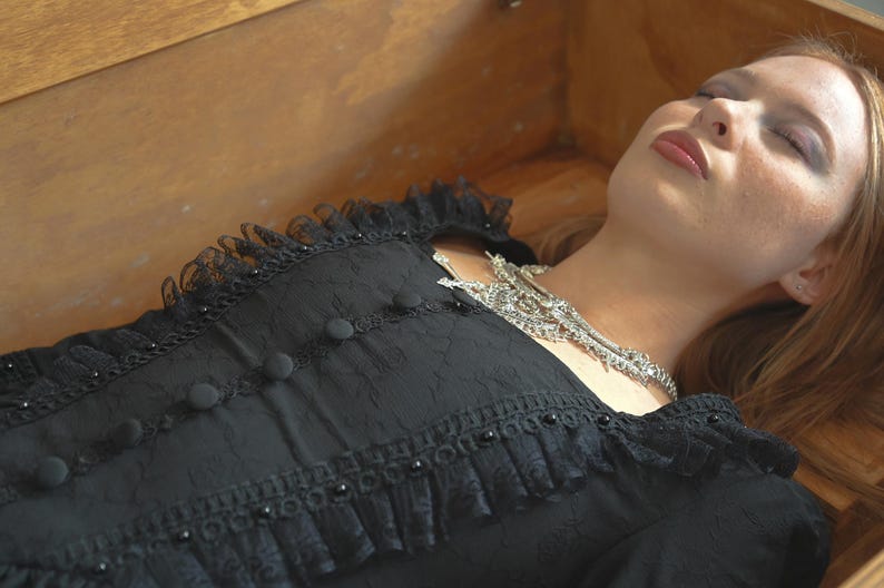 Op de afbeelding: Een vrouw ligt in een houten kist, gekleed in een zwarte kanten jurk met knopen en een zilveren ketting. Haar ogen zijn gesloten en haar mond staat lichtjes open.