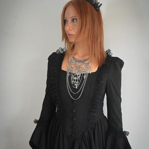 Op de afbeelding: Een vrouw in een zwarte jurk met kantdetails en een zilveren ketting met meerdere hangers. Ze draagt een zwarte kroon op haar hoofd en heeft rood haar.