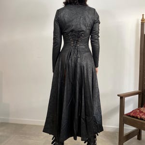 May include: A long black coat with a textured finish, featuring a lace-up back and a flared skirt. The coat has long sleeves and is paired with platform boots. The overall style suggests a gothic or alternative fashion aesthetic.