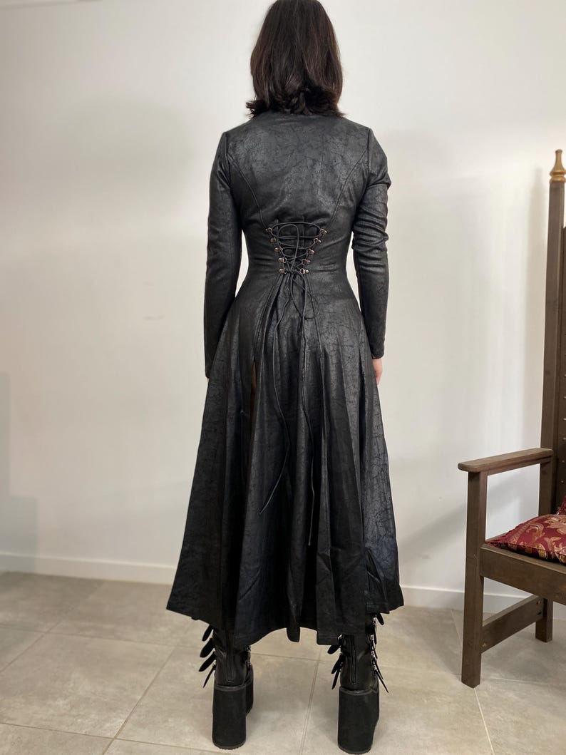 May include: A long black coat with a textured finish, featuring a lace-up back and a flared skirt. The coat has long sleeves and is paired with platform boots. The overall style suggests a gothic or alternative fashion aesthetic.
