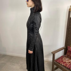 May include: A long black coat with a high collar, long sleeves, and a flared skirt. The coat features decorative straps and lacing details. The person is wearing black platform shoes and sunglasses.
