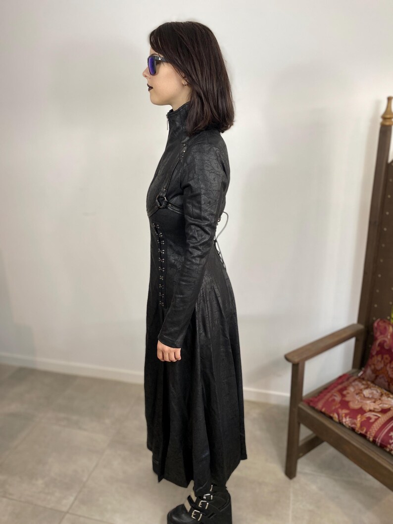May include: A long black coat with a high collar, long sleeves, and a flared skirt. The coat features decorative straps and lacing details. The person is wearing black platform shoes and sunglasses.