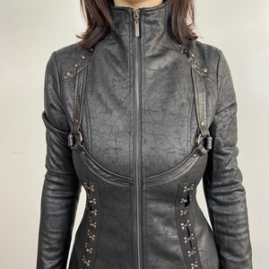 May include: Black faux leather jacket with a high collar and a full-length zipper. The jacket features decorative straps, metal accents, and a corset-style closure on the sides. The long-sleeved jacket has a textured finish.