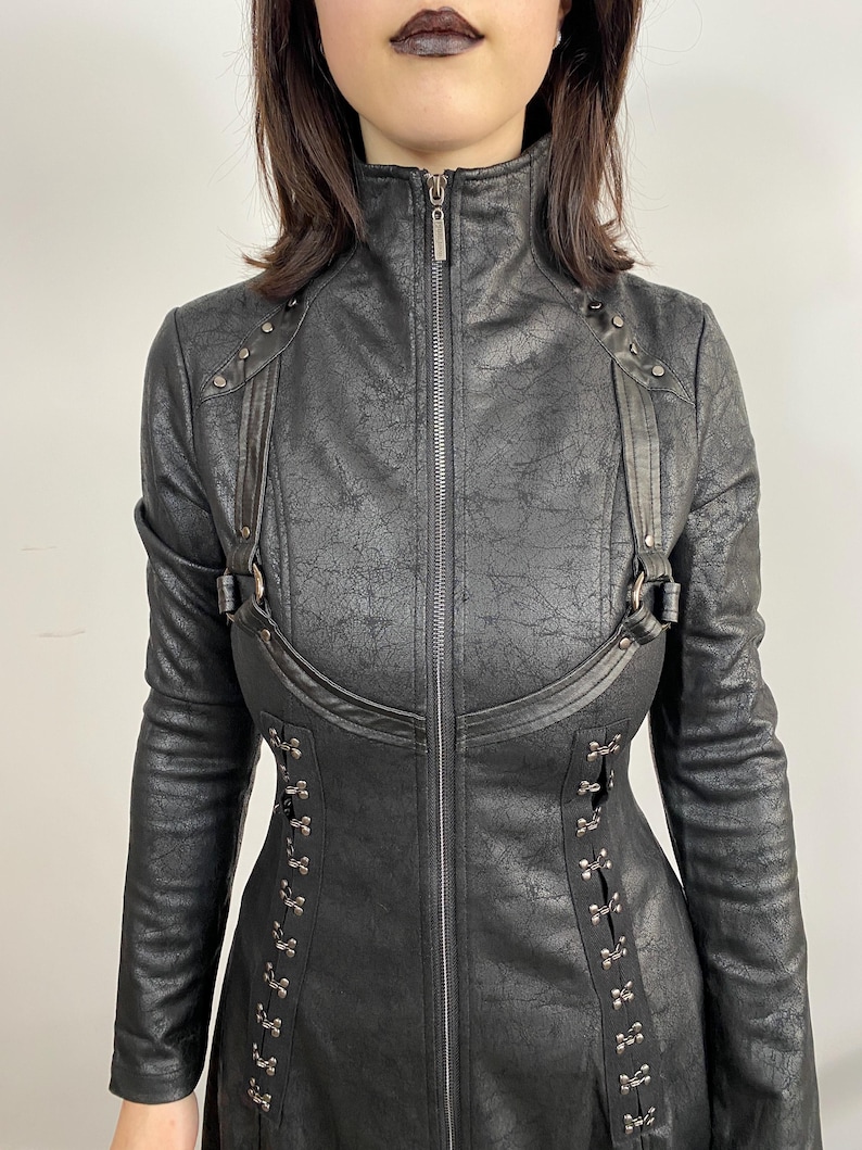 May include: Black faux leather jacket with a high collar and a full-length zipper. The jacket features decorative straps, metal accents, and a corset-style closure on the sides. The long-sleeved jacket has a textured finish.