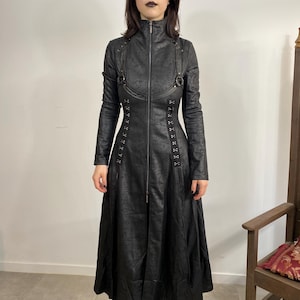 May include: A long, black coat with a textured pattern, a high collar, and a full-length zipper. The coat features decorative straps, grommets, and a flared skirt. The person in the image is wearing black boots and sunglasses.