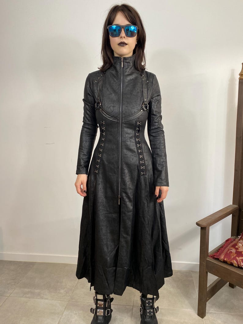 May include: A long, black coat with a textured pattern, a high collar, and a full-length zipper. The coat features decorative straps, grommets, and a flared skirt. The person in the image is wearing black boots and sunglasses.