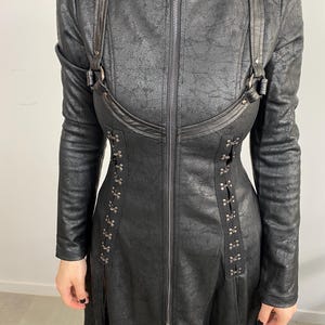 May include: Black faux leather coat with long sleeves, a high collar, and a full-length zipper. The coat features decorative straps, metal accents, and a textured finish, creating a gothic or steampunk aesthetic.