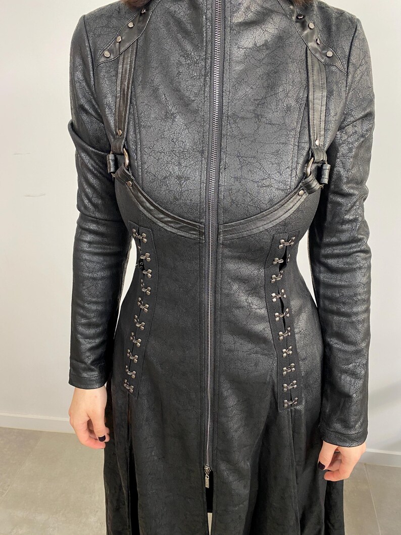 May include: Black faux leather coat with long sleeves, a high collar, and a full-length zipper. The coat features decorative straps, metal accents, and a textured finish, creating a gothic or steampunk aesthetic.