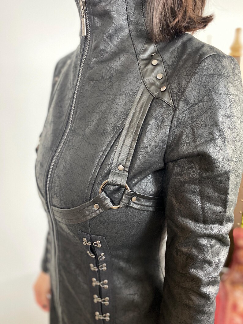 May include: A black faux leather jacket with a high collar and a textured finish. The jacket features a zipper closure, decorative metal hardware, and a unique harness-style detail with a metal ring.