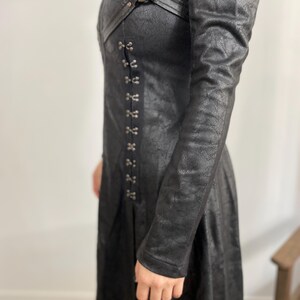 May include: A long black coat with a textured, leather-like appearance. The coat features a series of metal clasps down the front and decorative straps near the shoulders. The sleeves are long, and the coat appears to be fitted.