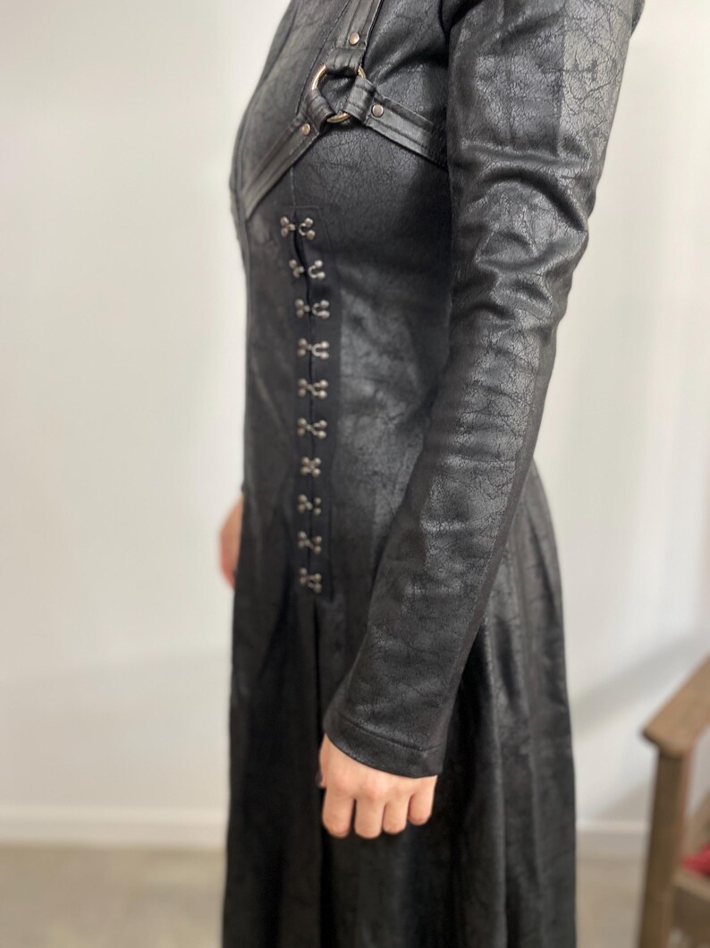 May include: A long black coat with a textured, leather-like appearance. The coat features a series of metal clasps down the front and decorative straps near the shoulders. The sleeves are long, and the coat appears to be fitted.