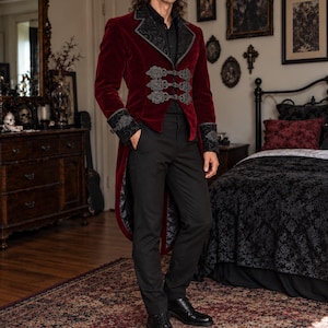 May include: A deep red velvet tailcoat featuring black ornate embroidered lapels and cuffs, paired with three decorative silver-toned metal clasps across the front. The garment is worn over a black button-down shirt and black trousers with black shoes.