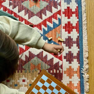 May include: A child plays a game of chess on a wooden board with a light blue and brown checkered pattern. The board is placed on a colorful rug with a geometric design.