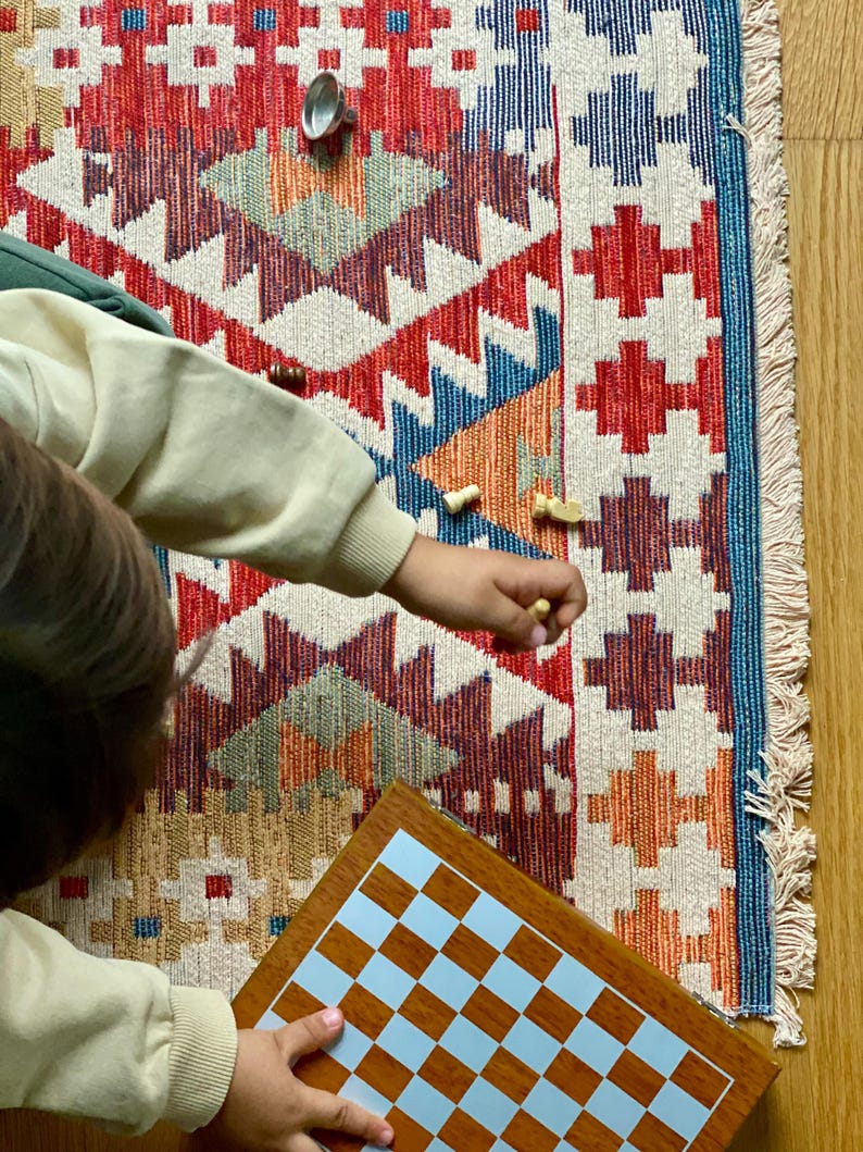 May include: A child plays a game of chess on a wooden board with a light blue and brown checkered pattern. The board is placed on a colorful rug with a geometric design.