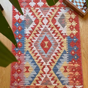 May include: A red, blue, and beige patterned rug with a geometric design. The rug has a fringe border.