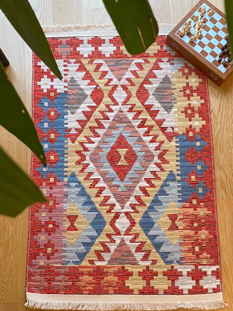 May include: A red, blue, and beige patterned rug with a geometric design. The rug has a fringe border.