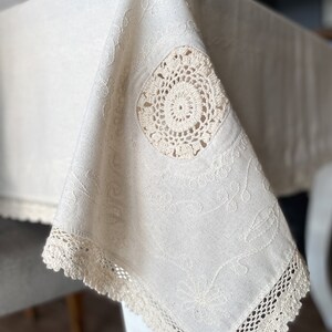 May include: A white linen tablecloth with delicate embroidery and crocheted trim. The tablecloth features a circular crocheted design in the center.