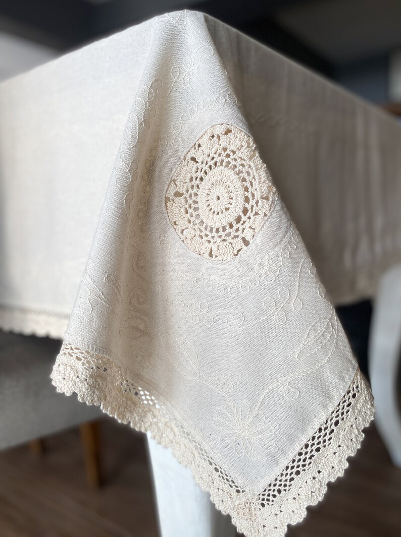 May include: A white linen tablecloth with delicate embroidery and crocheted trim. The tablecloth features a circular crocheted design in the center.