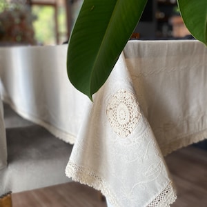 May include: Cream-coloured linen tablecloth with intricate embroidered details and a crocheted lace border. A circular crocheted doily is sewn onto the corner. The tablecloth is draped over a grey chair.